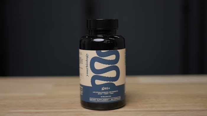 A bottle of gut+ by yourbiology probiotic supplement standing on a light brown wooden table with a black backdrop
